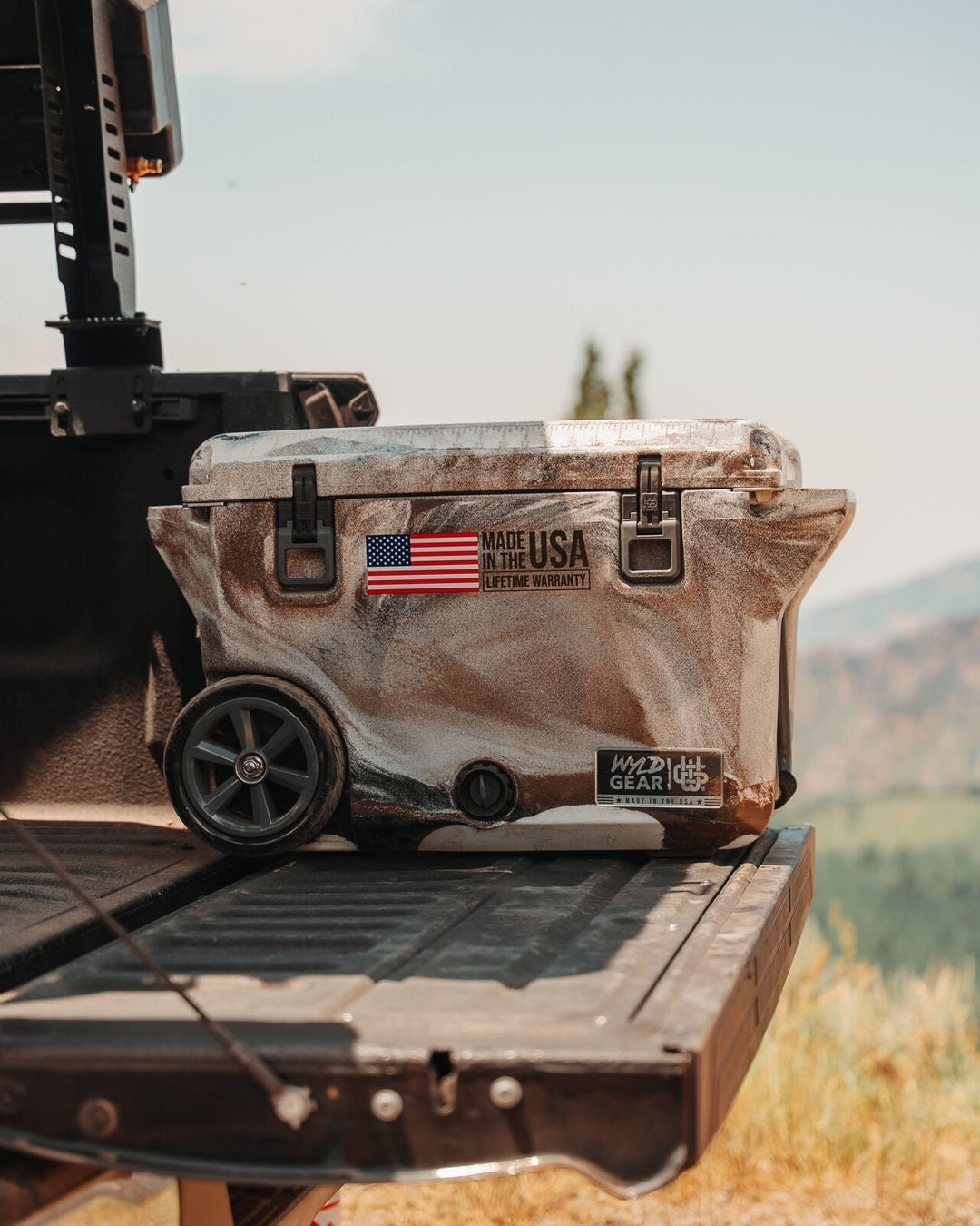 Wyld Gear Prairie cooler with wheels sitting on a truck tailgate in an open field, showing the Made in the USA lifetime warranty badge